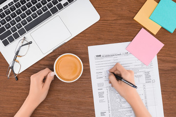 cropped view of woman holding coffee cup and filling tax form at workplace