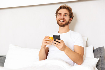 Handsome young bearded man in bed at home drinking juice using mobile phone.