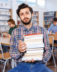 Tired man sitting with pile of books in university library on background with working fellow students