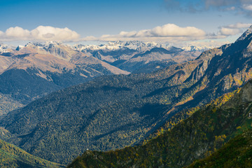 Naklejka premium Mountain landscape. Warm autumn Sunny day. The slopes of the mountains covered with autumn woods. Clouds above mountain peaks. The snow on the tops