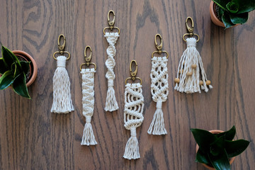 Five hand made cotton macrame key chains of different styles. Background is a beautiful wooden floor with green potted plants. - obrazy, fototapety, plakaty