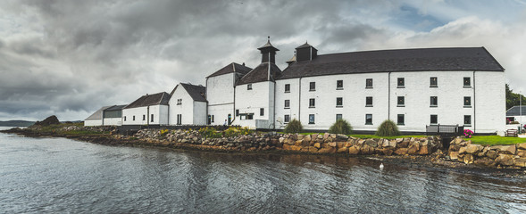 Panoramic view of Laphroaig distillery buildings. Islay island, Scotland. Grey cloudy sky before the rain. White cottages on the Scottish shoreline. Amazing autumn British landscape. - obrazy, fototapety, plakaty