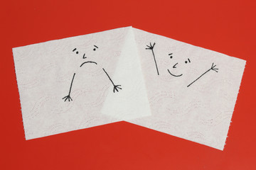 Two white toilet paper pieces sketched with a sad and a cheerful face, close to each others, representing the sadness and the happiness feelings
