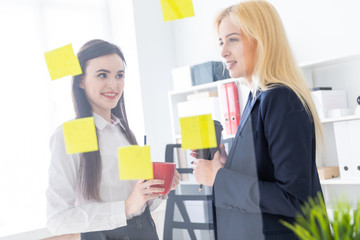 Two girls talking in the office. Girls are a dialogue near a transparent Board with stickers.