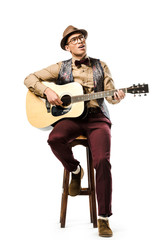 smiling mixed race male musician in hat and eyeglasses playing on acoustic guitar while sitting on chair isolated on white