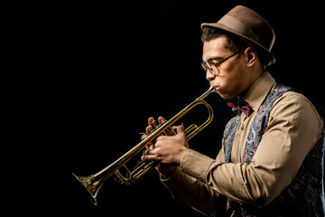 side view of mixed race male jazzman in hat and eyeglasses playing on trumpet isolated on black