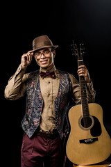 cheerful mixed race male musician in eyeglasses and hat posing with acoustic guitar isolated on black