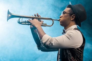 side view of jazzman playing on trumpet on stage with dramatic lighting and smoke