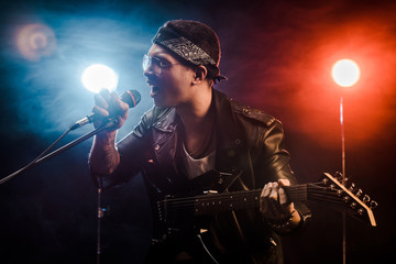 emotional male musician singing in microphone and playing on electric guitar on stage during rock concert