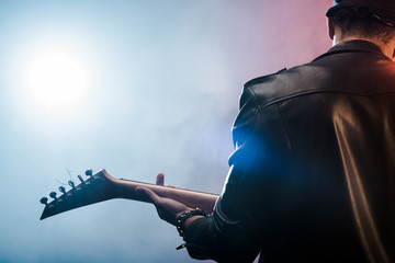 rear view of male rock star in leather jacket performing on electric guitar on stage with smoke and...