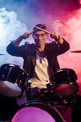 happy mixed race male musician raising drum sticks while sitting behind drum set on stage with dramatic lighting and smoke