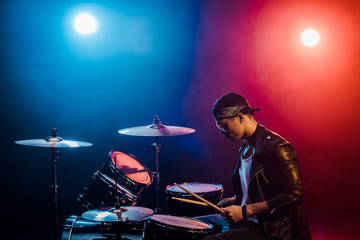 male musician in leather jacket playing drums during rock concert on stage with smoke and spotlights