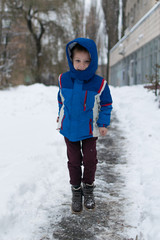 A little boy in a blue winter jacket is jumping in the street with snow.
