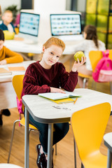 adorable schoolkid holding apple and looking at camera while sitting with classmates in library