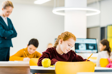 teacher looking at adorable schoolchildren writing and studying at desks