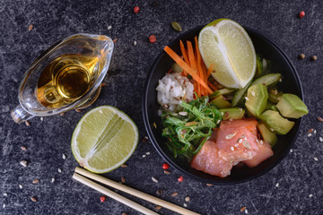 Homemade healthy Poké bowl with rice, salmon, avocado, white onion, scallion greens, sesame oil, soy and sesame sauce, seaweed (wakame), lime and rice vinegard on a dark backgound