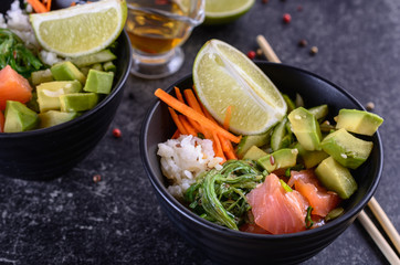 Homemade healthy Poké bowl with rice, salmon, avocado, white onion, scallion greens, sesame oil, soy and sesame sauce, seaweed (wakame) and other ingredients in a black bowl on a dark backgound