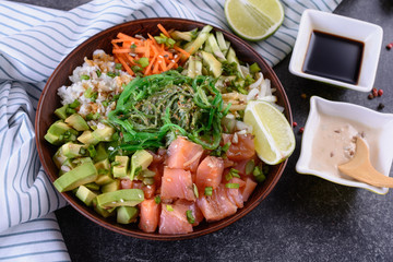 Homemade healthy Poké bowl with rice, salmon, avocado, white onion, scallion greens, sesame oil, soy and sesame sauce, seaweed (wakame) and other ingredients on a dark backgound, top view, close up