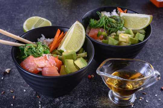 Two homemade healthy Pok&eacute; bowl with rice, salmon, avocado, white onion, scallion greens, sesame oil, soy and sesame sauce, seaweed (wakame), lime and rice vinegard on a dark backgound
