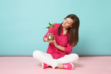 Full length of woman in rose shirt, white pants sitting on floor hold gold coins in glass jar isolated on bright pink blue pastel wall background studio. Fashion lifestyle concept. Mock up copy space.