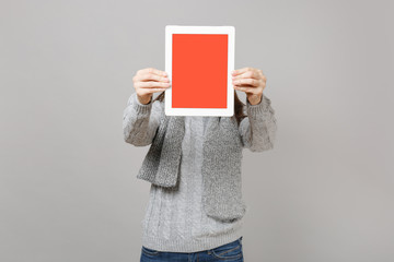 Young woman in gray sweater, scarf hiding, covering face with tablet pc computer with blank empty screen isolated on grey background. Healthy lifestyle online treatment consulting cold season concept.