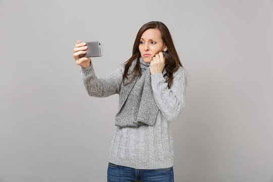 Upset young woman in sweater, scarf doing selfie shot on mobile phone, making video call isolated on grey background. Healthy fashion lifestyle people emotions cold season concept. Mock up copy space.