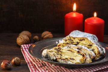 christmas cake, german christstollen with fruits, raisins and marzipan in front of two red candles and nuts on rustic dark wood