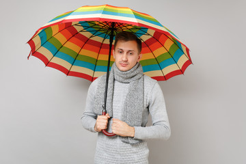 Pensive young man in gray sweater, scarf looking down, holding colorful umbrella isolated on grey background. Healthy fashion lifestyle people sincere emotions cold season concept. Mock up copy space.