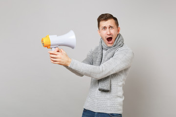 Crazy young man in gray sweater, scarf holding megaphone keeping mouth wide open isolated on grey background. Healthy fashion lifestyle people sincere emotions cold season concept. Mock up copy space.