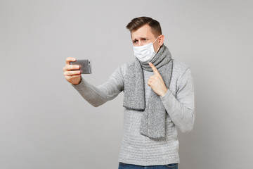 Young man in sweater, scarf sterile face mask making video call with mobile phone, pointing index finger on himself isolated on grey background. Health ill sick disease treatment, cold season concept.