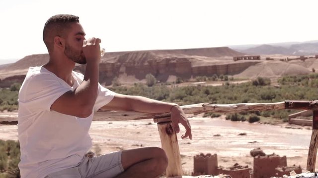 A Moroccan guy drinking tea on a sunny day above the Kasbah of Ait Benhaddou