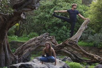 Young couple in urban clothing performing yoga asanas on top of tree trunk. Smiling woman in utpluthih and man in utthita hasta padangusthasana
