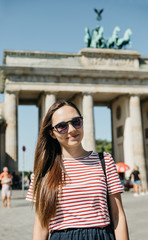 Obraz premium Portrait of a young beautiful positive smiling stylish tourist girl in Berlin in Germany. Brandenburg Gate and unrecognizable blurred people in the background.