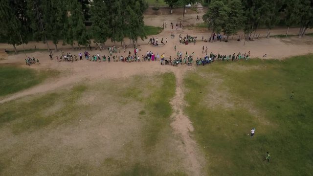 Aerial View of a Large Group of African Students Gathered to Watch Their Students and American Visitors Play a Game of Soccer on a Dirt and Grass Field, Rotating and Panning Motion