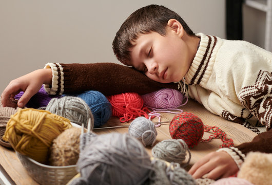 Child boy is learning to knit. He is dreaming and closing eyes. Colorful wool yarns are on the wooden table.
