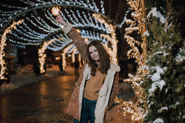 Pretty dark haired girl wearing blue jeans and beige top with snowflakes Christmas lights outdoor at night time.