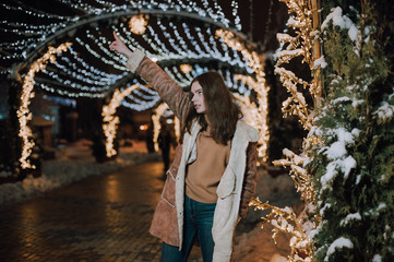 Pretty dark haired girl wearing blue jeans and beige top with snowflakes Christmas lights outdoor at night time.