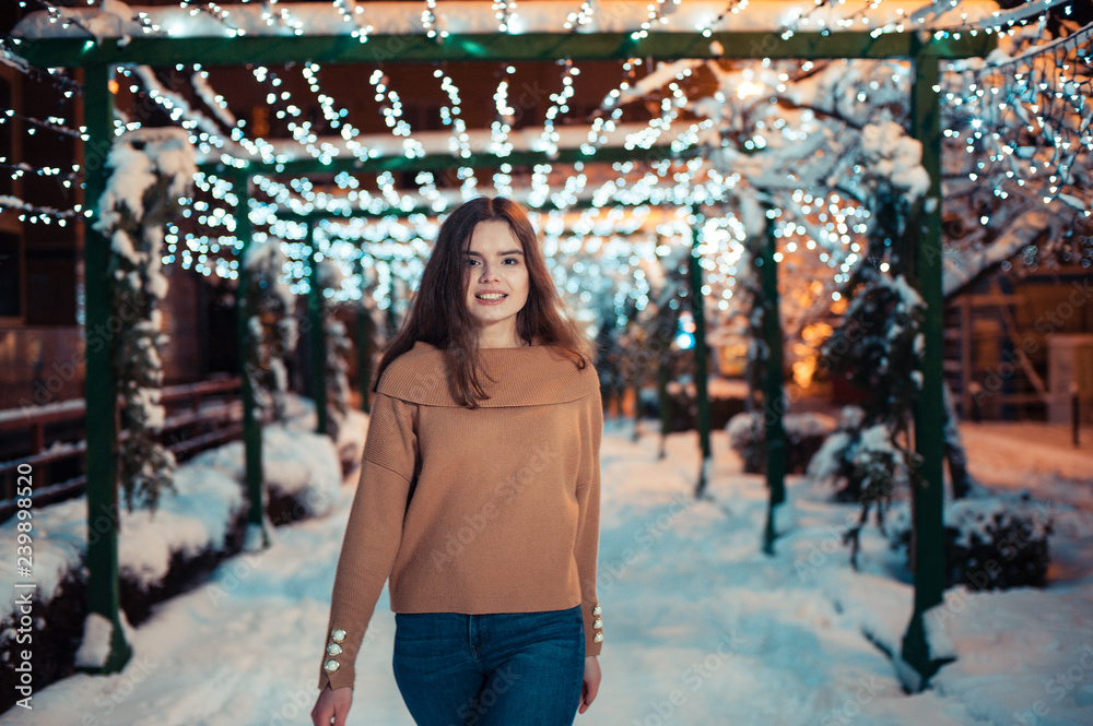 Wall mural Pretty dark haired girl wearing blue jeans and beige top with snowflakes Christmas lights outdoor at night time.