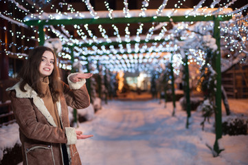 Pretty dark haired girl wearing blue jeans and beige top with snowflakes Christmas lights outdoor at night time.