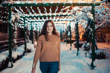 Fototapeta na wymiar Pretty dark haired girl wearing blue jeans and beige top with snowflakes Christmas lights outdoor at night time.