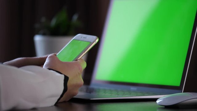 Holding a smartphone with green screen, scrolling and then clicking with left finger on the upper left corner of the screen with a laptop with green screen in the back