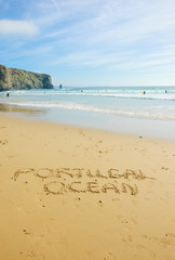 Words PORTUGAL and OCEAN are written on the scenic beach in the Algarve region of Portugal. People swimming and surfing.