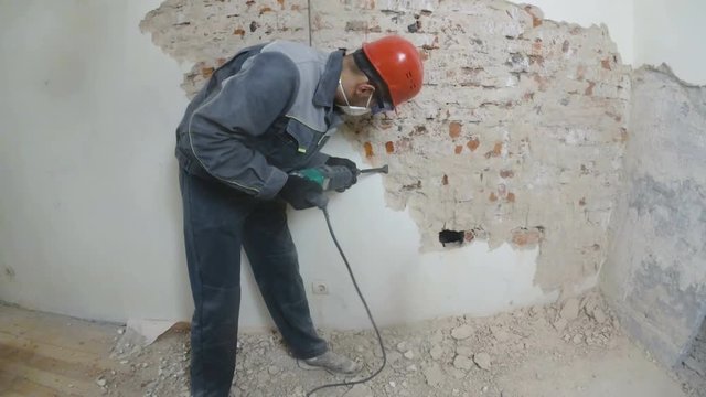 worker in protective suit demolishes plaster wall. Dirty, hard work. Personal protective equipment. Helmet, respirator and goggles.