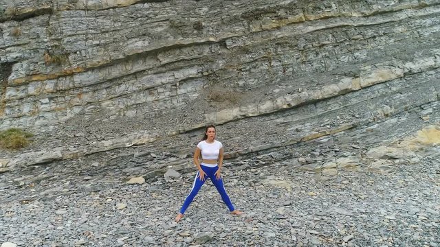 Woman is doing squats the bodyflex during breathing exercises on the rock background. Aerial beautiful footage view.