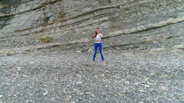 Woman is doing squats the bodyflex on the rock and sea background. Aerial beautiful footage view.