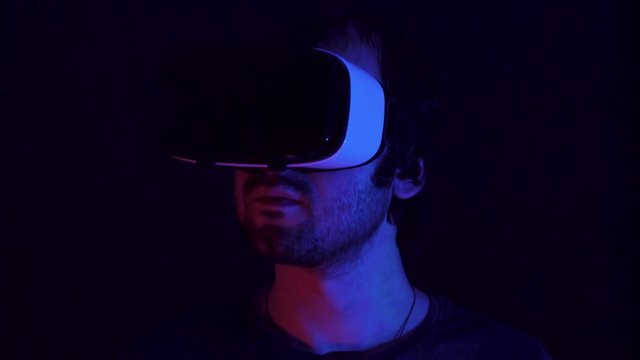 Young man in VR headset looks around and wonders how amazing. Virtual reality helmet on black technology background. Modern fluorescent light
