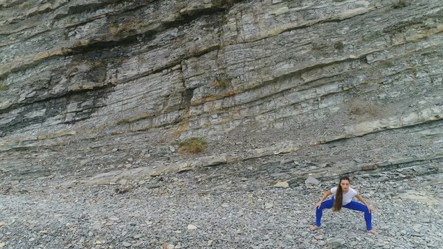 Woman is doing plie squats the bodyflex during breathing exercises on the rock background. Aerial beautiful footage view.