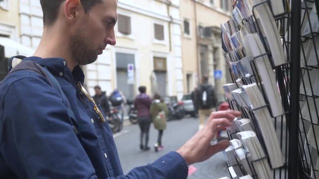 Male Backpacker Picking Out A Postcard On A Busy Tourist Street In Rome.
