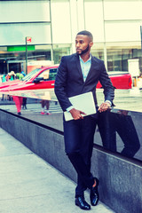 Obraz premium Young African American Businessman with beard, afro short hair, traveling in New York, wearing black suit, leather shoe, holding laptop computer, standing on street in Middletown of Manhattan, looking