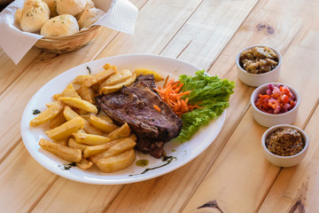 Plato blanco de comida casera bife de carne con papas fritas ,ensalada, condimentos , pan sobre mesa de madera con luz natural